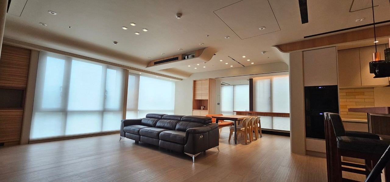 客餐廳公領域採現代簡約風格，以暖木色地坪搭配白色牆面。天花板施作弧形降板修飾空調風口，結合大面採光風琴簾與俐落皮質沙發，型塑開闊流暢的開放格局。
