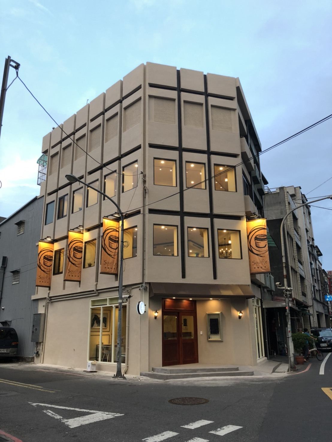 商業空間建築外觀以米黃色塗料搭配深色格狀立面線條，轉角入口規劃木質大門與落地窗，二至三樓設置規律排列的方格觀景窗，形成對稱明亮的街角立面。