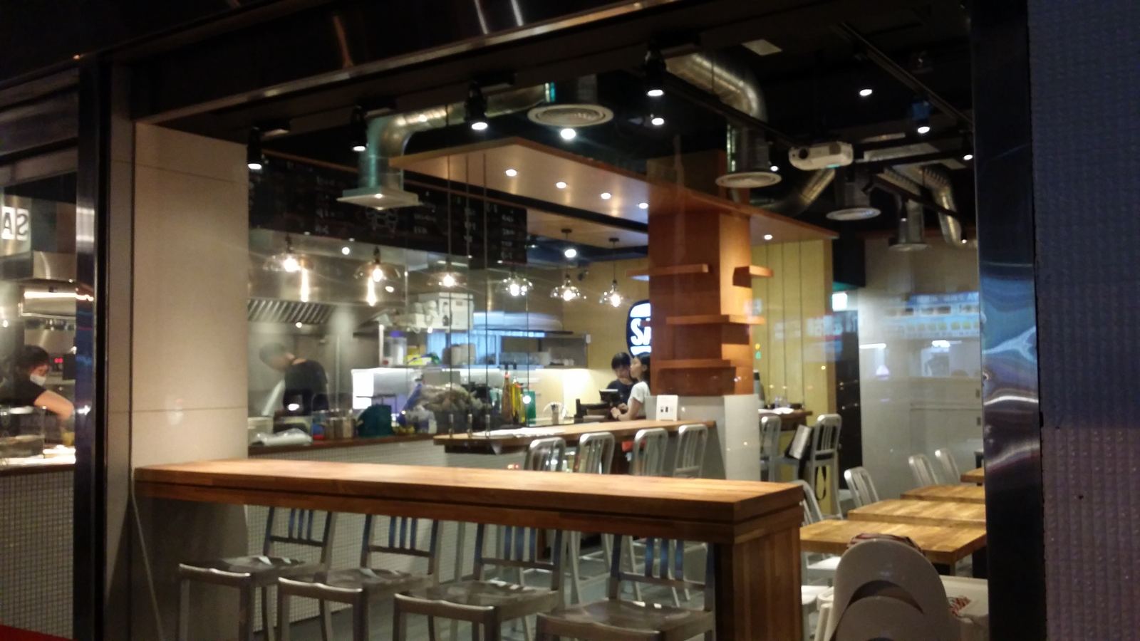 商業餐飲空間呈現工業風格，以天花板裸露的金屬風管搭配深色天花與軌道投射燈，結合原木吧台與金屬高腳椅，配置開放式廚房格局。