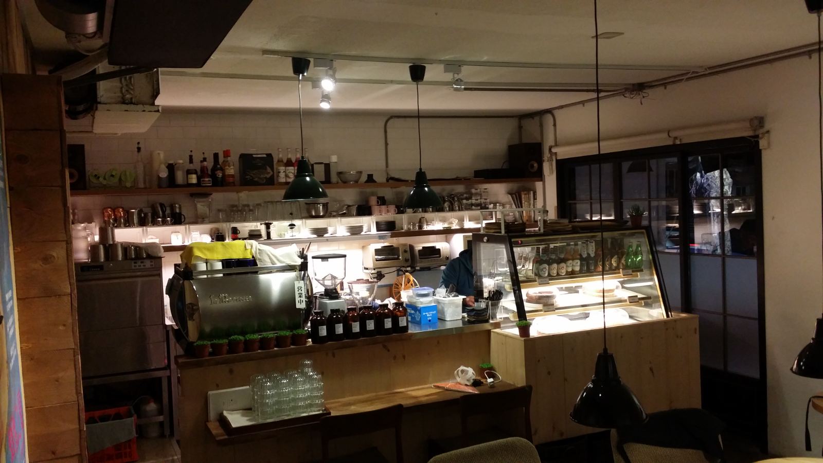 商業咖啡空間採用溫潤木質吧台搭配不鏽鋼工作區，天花板施作裸露明管與復古金屬吊燈，並以背牆開放層架滿足收納與陳列機能。