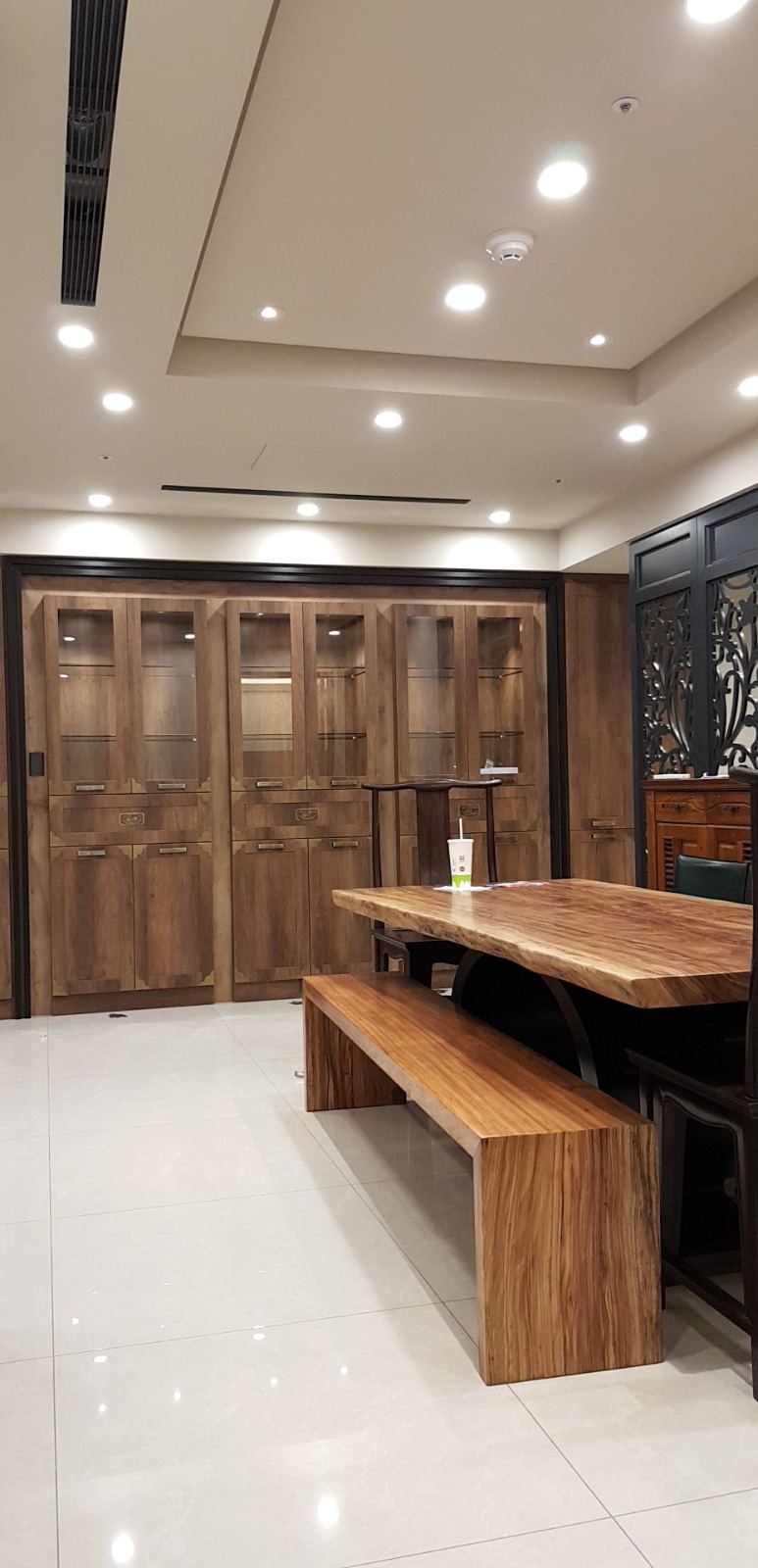 用餐與品茗空間以厚實原木長桌搭配實木長凳為核心，背牆規劃整片帶有金屬飾件的深木色玻璃展示櫃，天花板則結合嵌燈照明與造型線條。