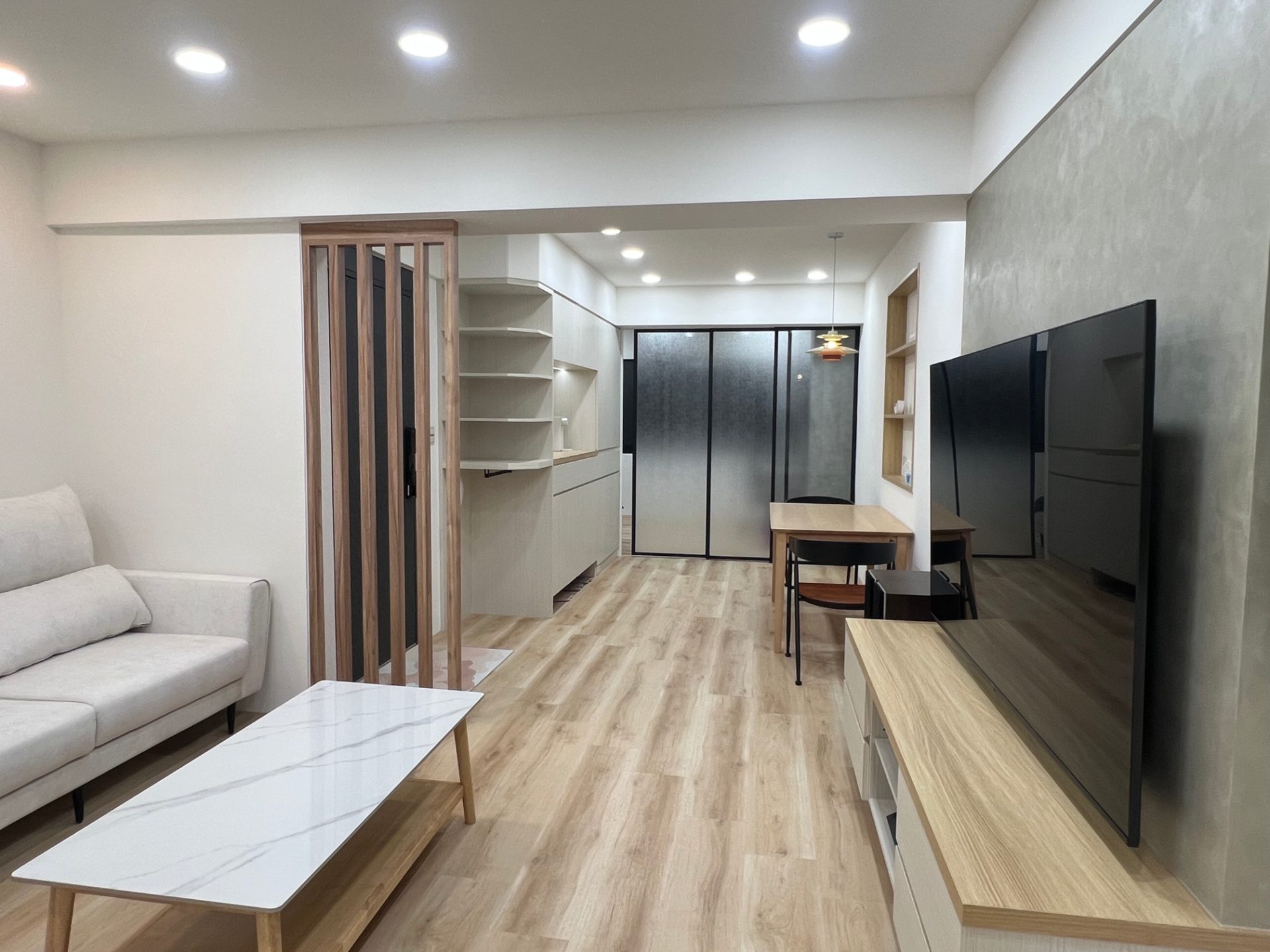 客廳與餐廚公領域展現日系無印與北歐自然風格，以淺木質地板鋪陳暖調基底，結合玄關木格柵屏風區隔場域，並運用黑框霧面玻璃拉門保持空間開闊感。