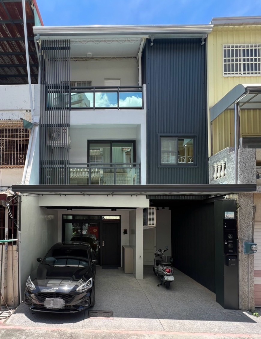 建築外觀與車庫展現現代簡約風格，整體以黑白對比色調鋪陳，立面結合深色金屬浪板、垂直遮陽格柵與透光玻璃欄杆，構築俐落分明的幾何層次。