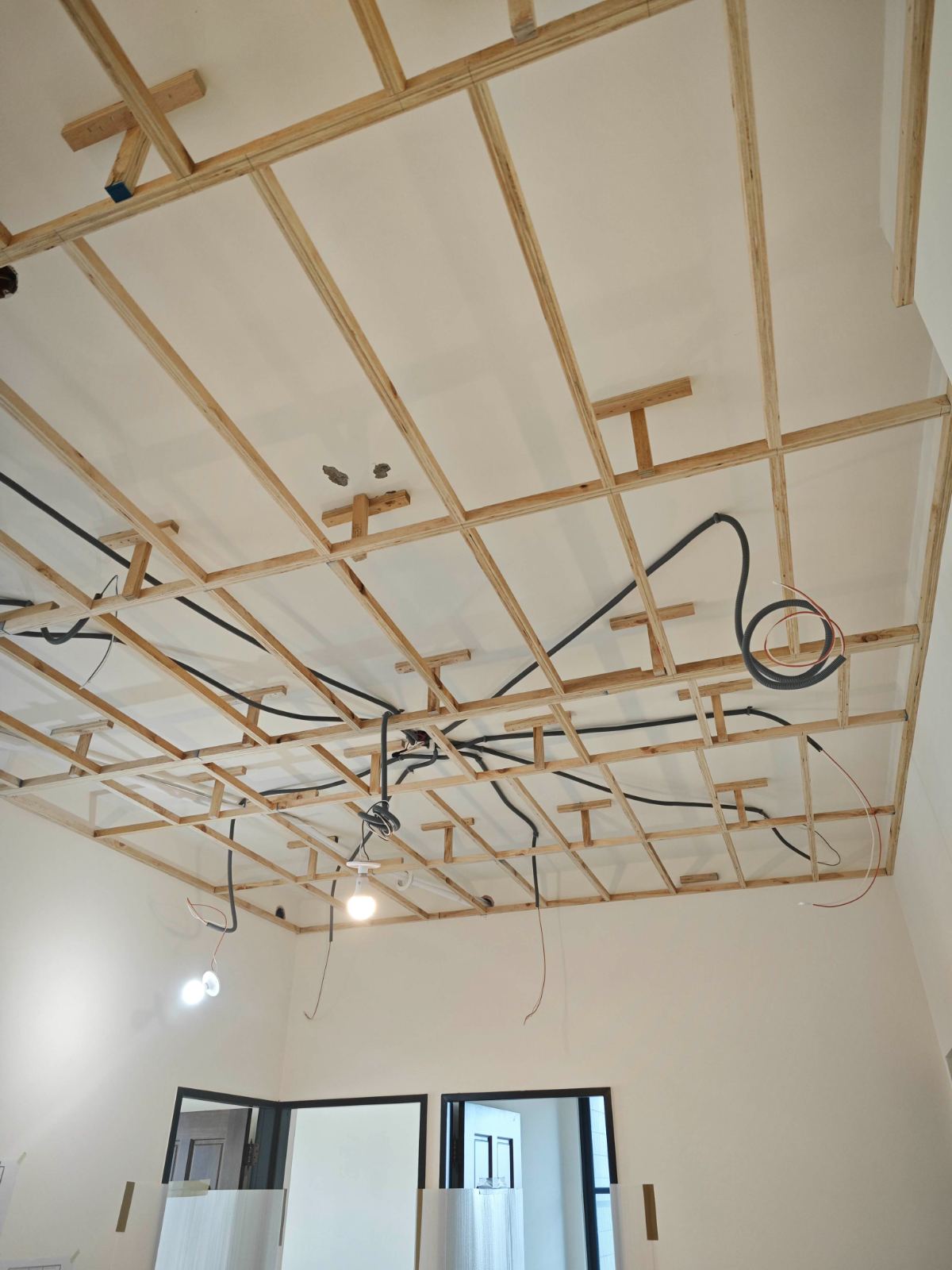 室內處於天花板木作工程施工階段，頂部架設格狀木角材骨架，並配置黑色電線波浪管與簡易施工照明燈具。