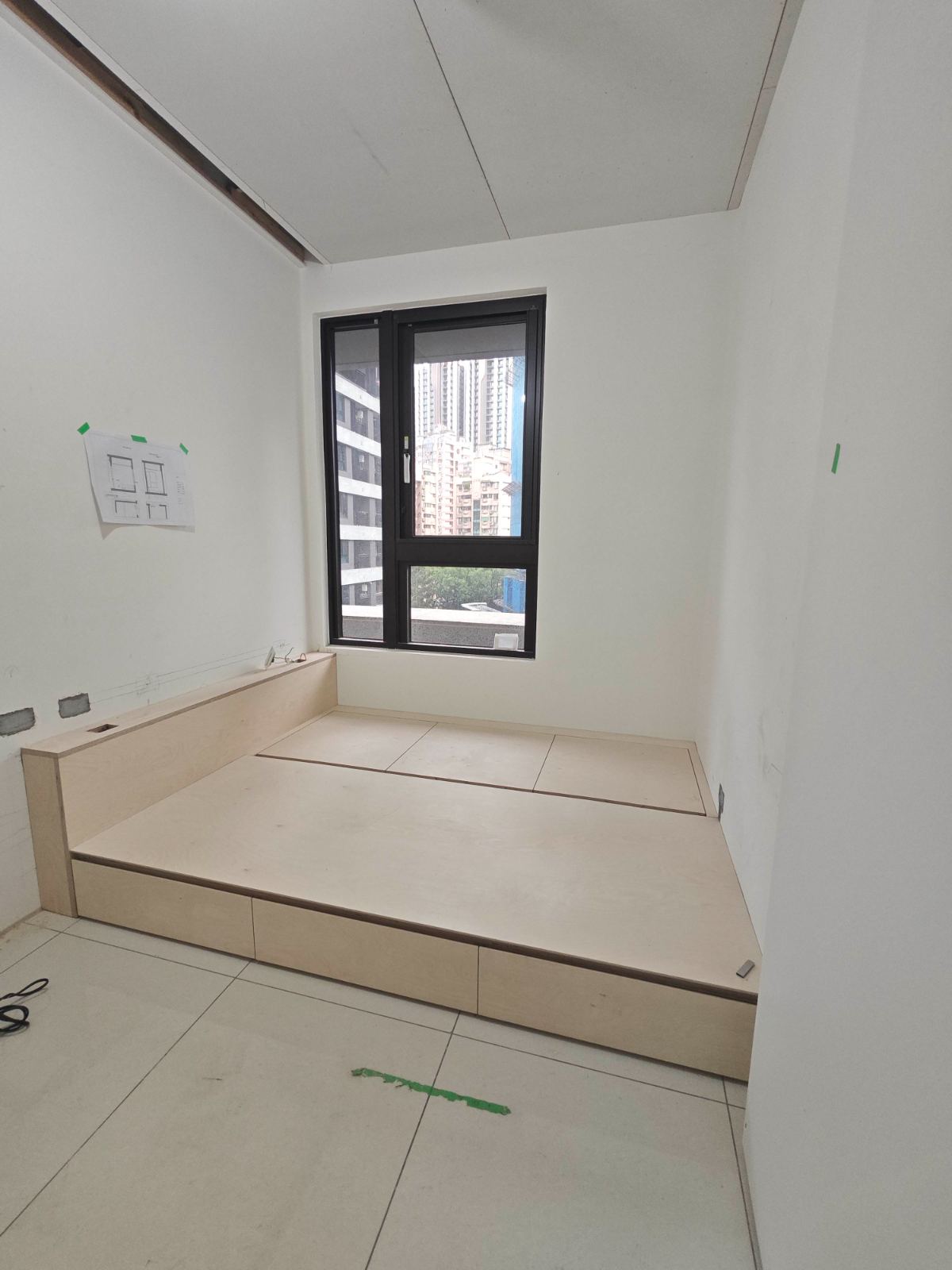 施工中的臥室空間以白色牆面搭配淺木色木作架高地台，地台整合側邊抽屜與上掀式收納機能，緊鄰黑色鋁框採光窗。