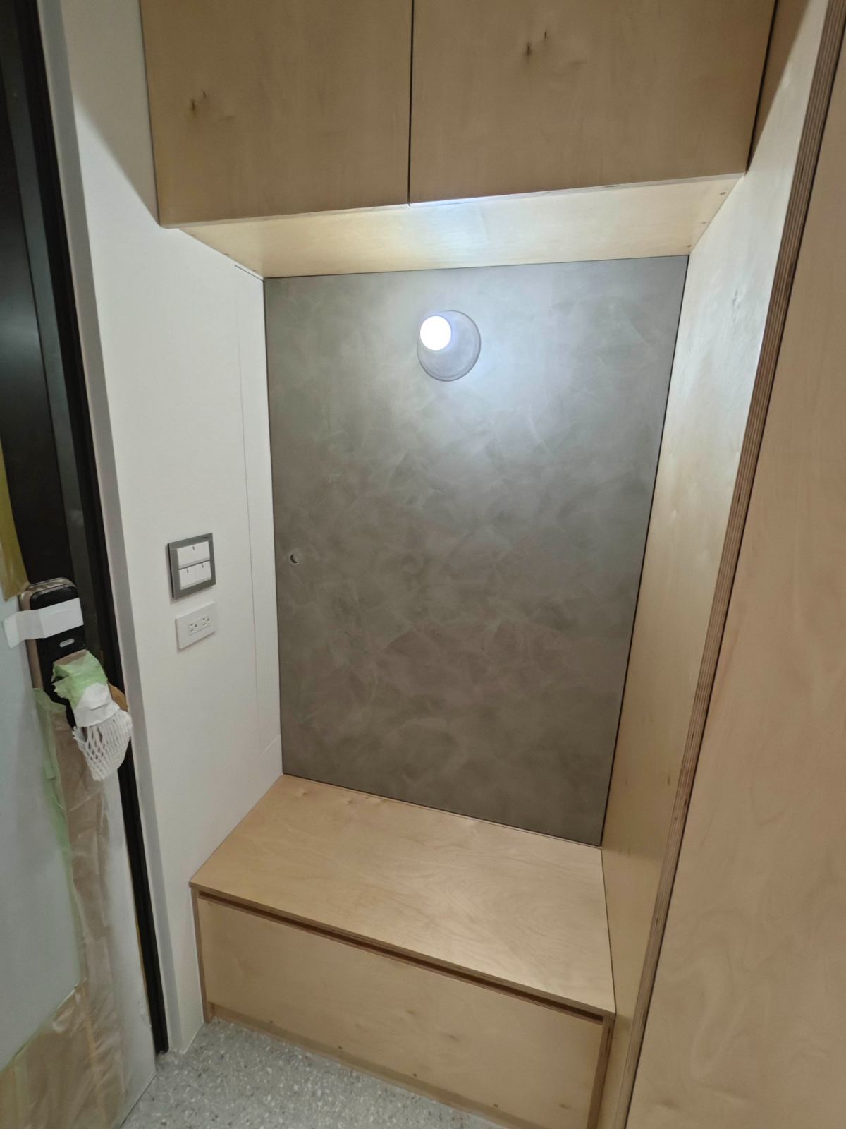 玄關空間以現代簡約風格呈現，運用淺色合板打造一體成型的收納櫃與下嵌穿鞋椅，壁面襯以灰色手刷塗料與極簡圓形壁燈點綴光影。