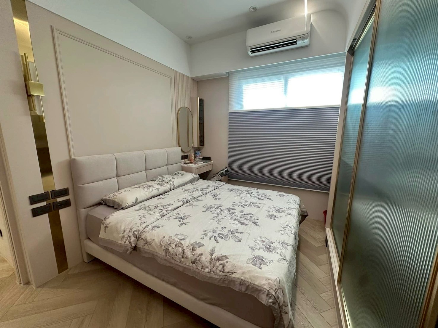 臥室鋪設人字拼木地板，床頭背牆結合細緻框線線板與金屬鏡面飾板，搭配米色方格軟包床頭與轉角梳妝區，側邊配置長虹玻璃推拉門，呈現典雅精緻的休憩空間。