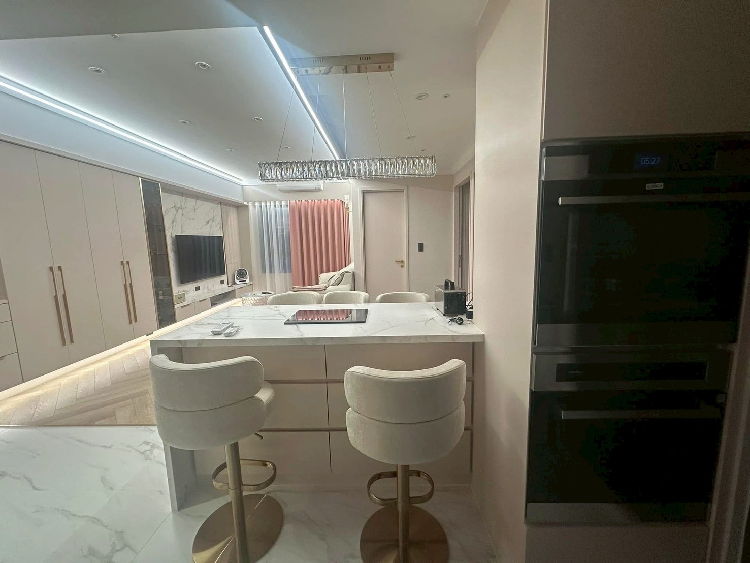 現代簡約風格的開放式客餐廚空間，以米白與柔和暖色為基底。天花板配置俐落線性燈帶，搭配大理石紋中島吧台與整面平整收納高櫃，呈現整潔連貫的空間視野。