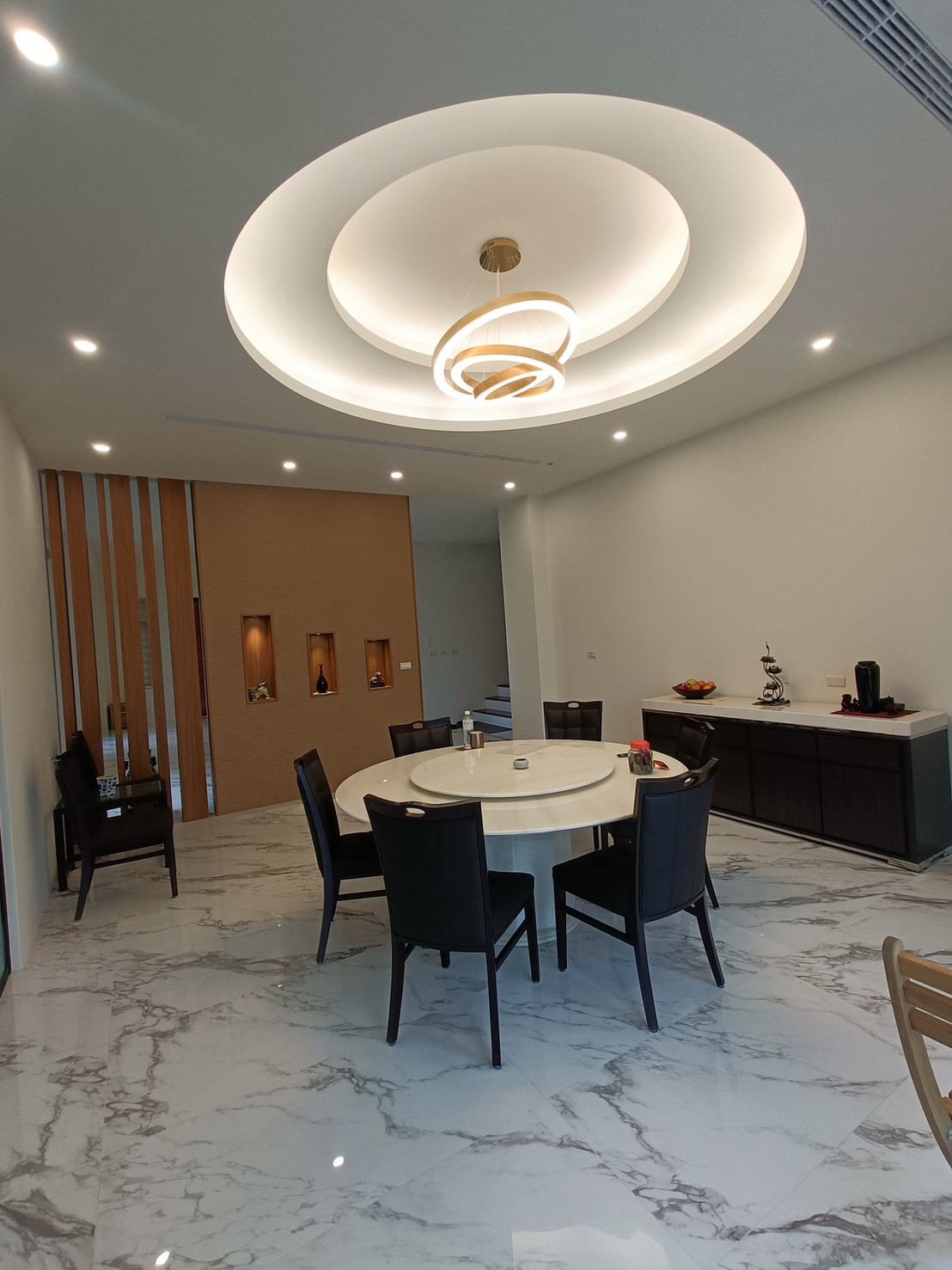 現代簡約風格的餐廳空間鋪設白色大理石紋地磚，天花板採多層次圓形凹槽並懸掛環形吊燈，背景立面結合木格柵與發光壁龕作為視覺分區。