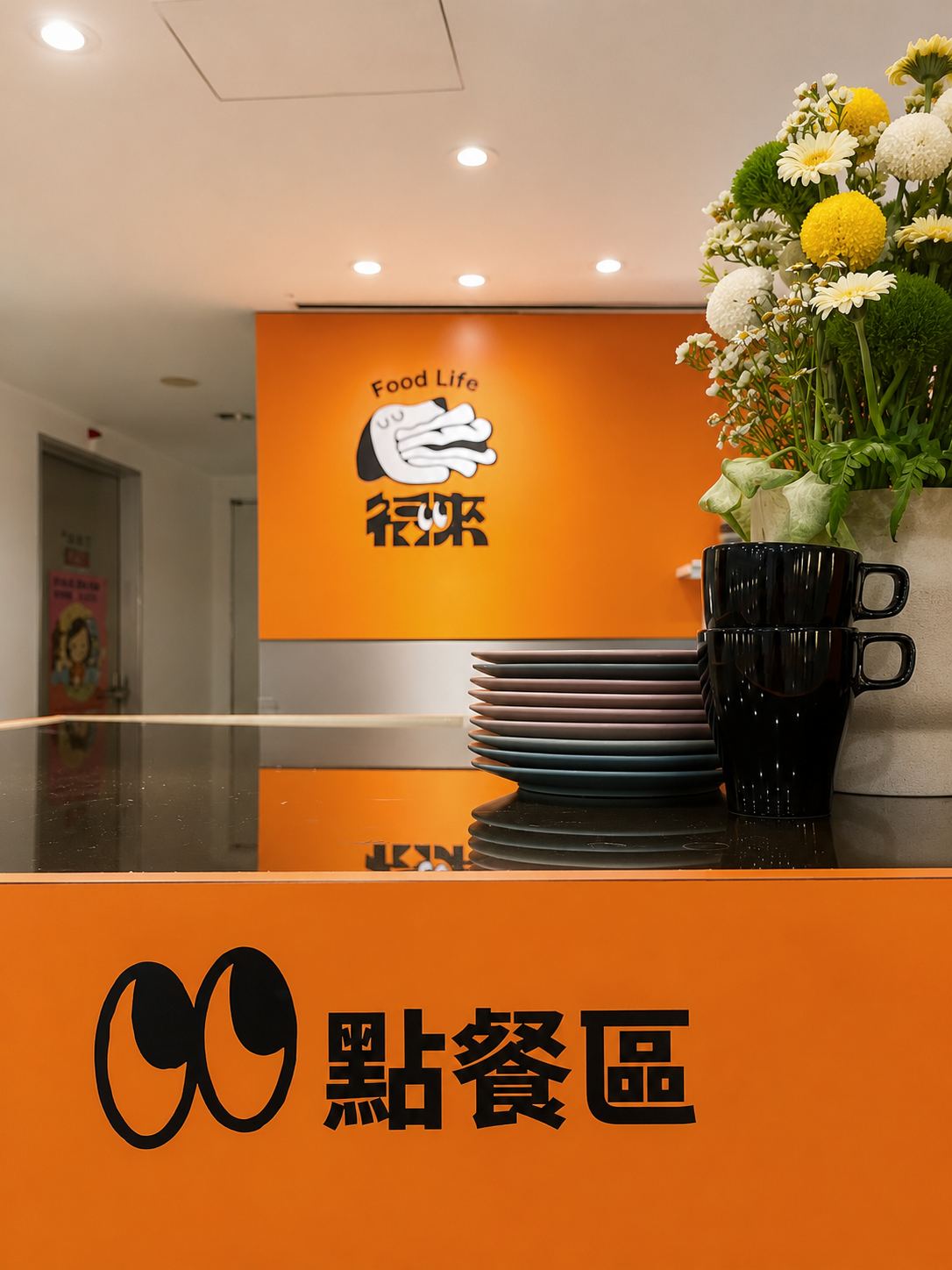 商業空間以鮮明亮橘色與白色為主調，櫃檯配置黑色亮面檯面與花藝擺設，背景牆結合品牌識別圖樣，並以天花板嵌燈均勻洗亮整體點餐區。